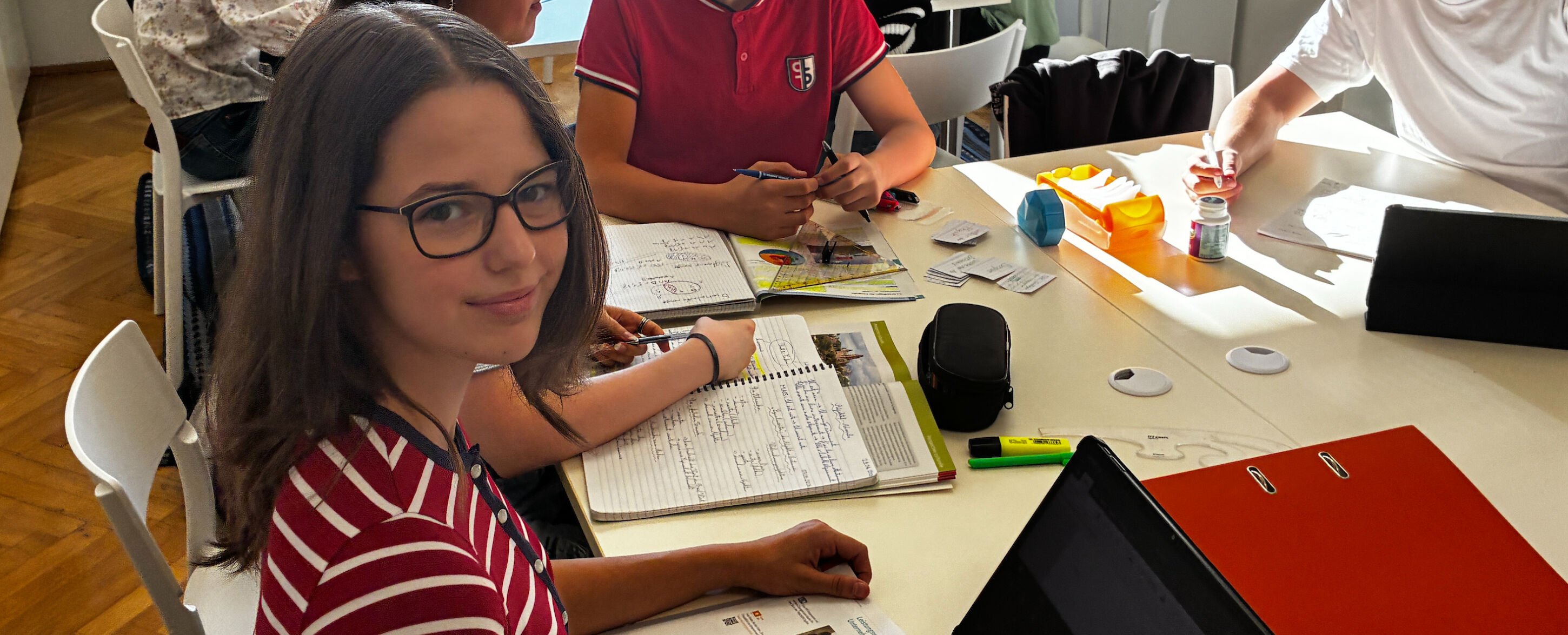 Jugendliche arbeiten gemeinsam an einem großen Tisch mit Schulbüchern, Heften und digitalen Geräten in einem hellen Lernraum.